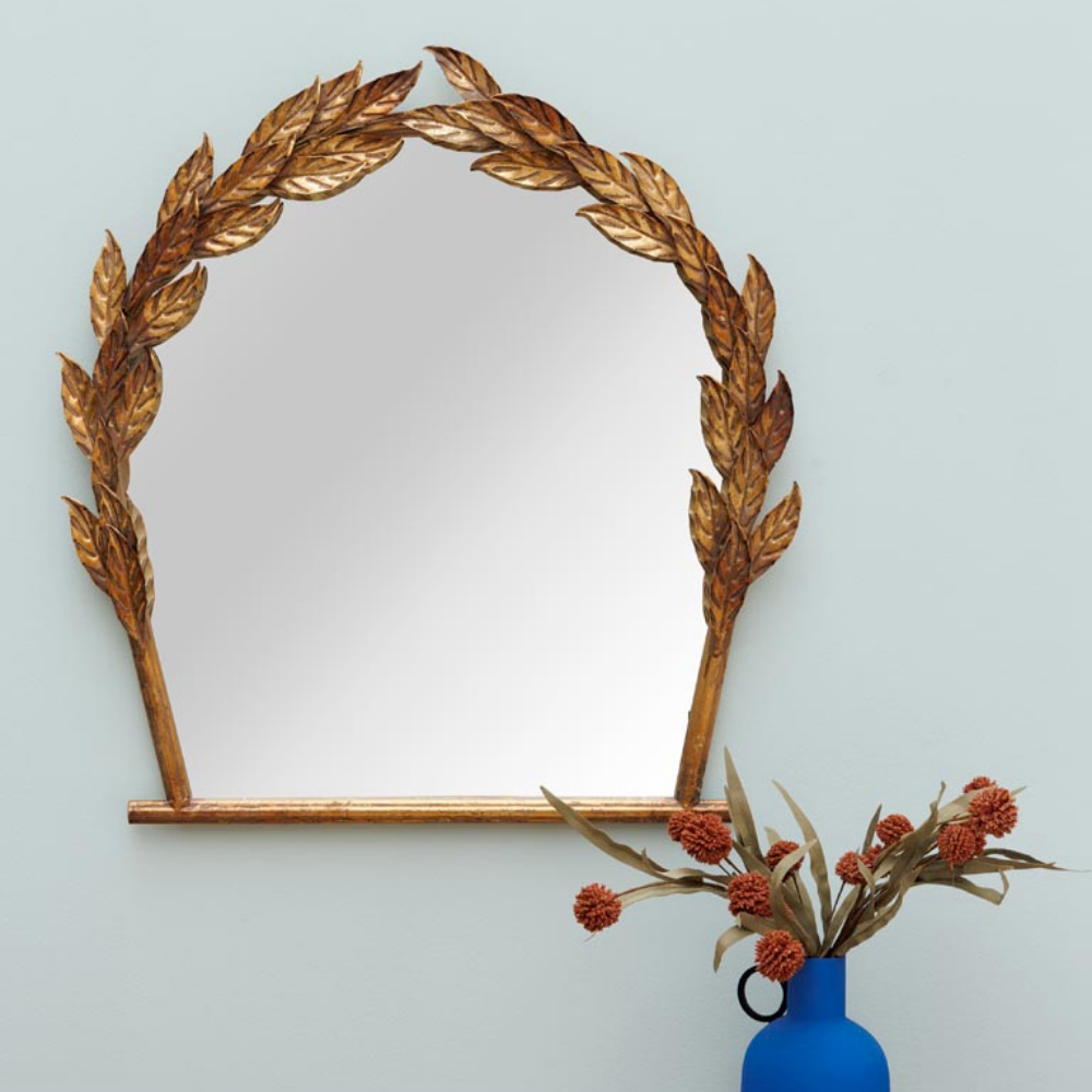 An arched wall mirror with a decorative gold leaf laurel frame, mounted on a light blue wall above a blue vase with dried orange flowers.