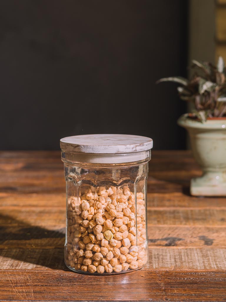 A clear glass jar with a light wood lid, filled with beige chickpeas, set on a rustic wooden table with a dark background and a blurred potted plant.