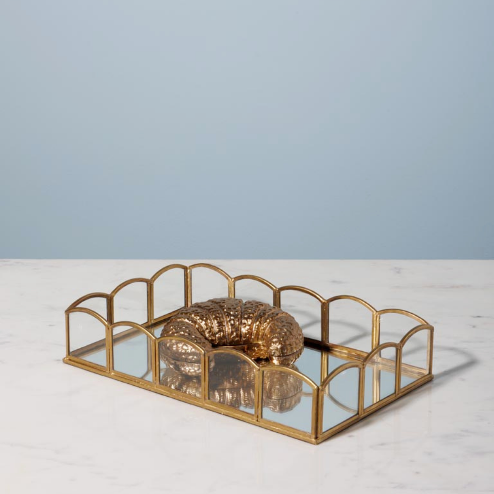 A rectangular gold-framed tray with mirrored glass panels and arched top details, holding a textured metallic gold decorative object, set on a white marble surface against a light blue background.