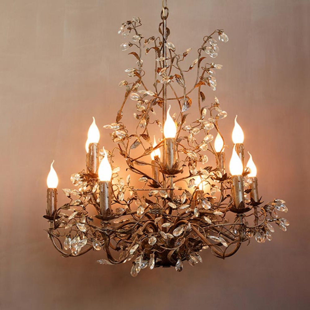 A multi-tiered chandelier with a bronze metal frame decorated with leaf and vine motifs, clear hanging crystal prisms, and multiple lit candle-style bulbs, shown against a warm beige wall.