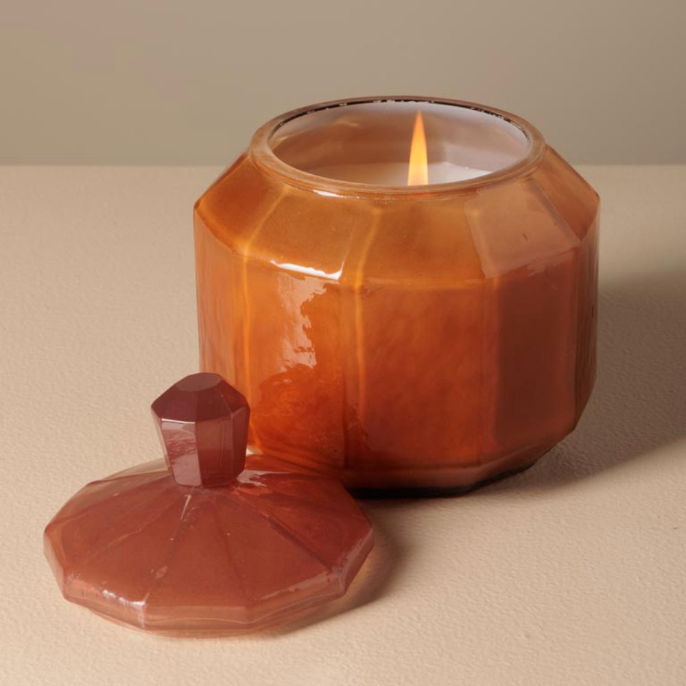 A faceted amber glass jar candle with a matching lid placed beside it on a neutral beige surface. The candle is lit, with the flame visible inside the glass container.