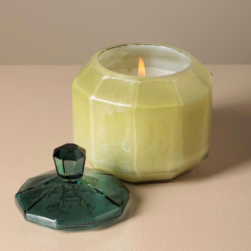 A faceted glass candle jar with a lit white candle inside, shown beside a matching faceted glass lid in dark green on a beige surface and background.