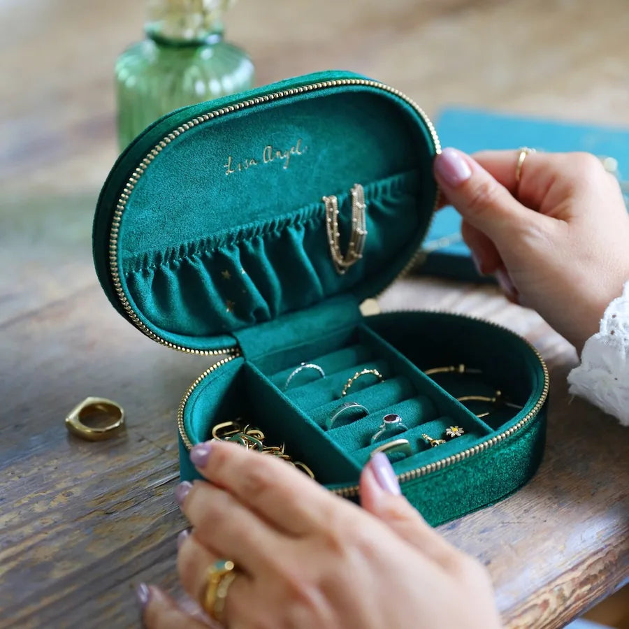 An open round green velvet jewelry box with gold-tone zippers and ring slots, shown on a wooden table with hands holding it and jewelry inside.