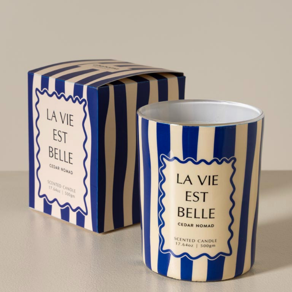 A scented candle in a cylindrical tin with blue and cream vertical stripes, shown next to its matching striped box. The label reads 'LA VIE EST BELLE' and 'CEDAR NOMAD'.