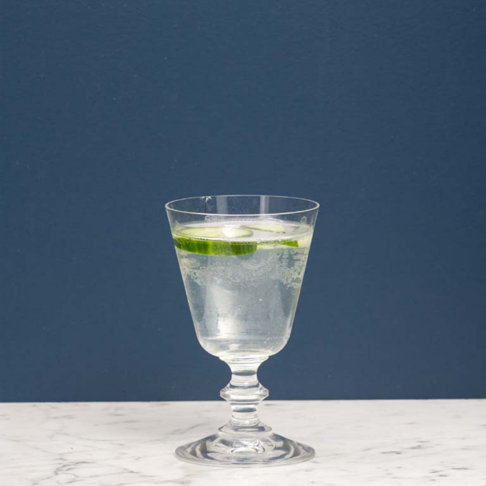 A clear glass goblet with a flared base, filled with a transparent bubbly drink and garnished with a lime slice, set on a white marble surface against a dark blue background.