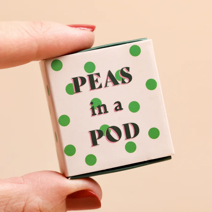 Small matchbox with 'Peas in a Pod' text and green polka dots held by a hand against a beige background