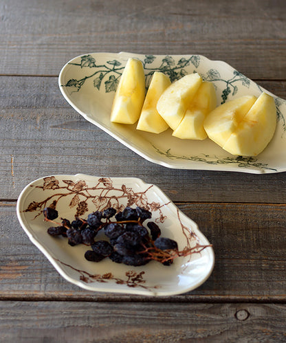 White ceramic oval-shaped plates with hand-painted botanical designs in green and brown, holding sliced apples and grapes on rustic wooden surface.