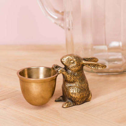 A small decorative rabbit figurine in an antique brass finish holding a matching metal cup, placed on a light wood surface with a soft pink background and a blurred glass vessel behind it.