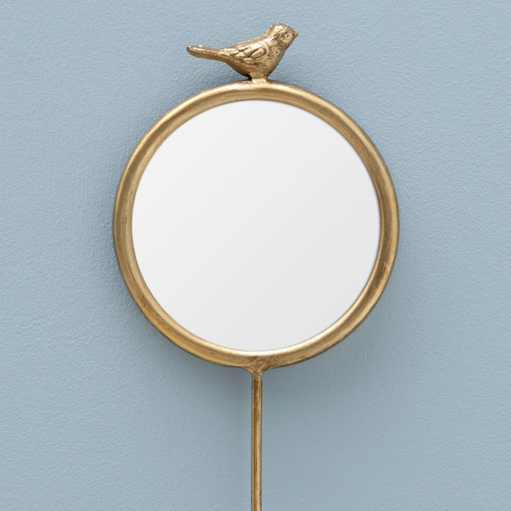 A round wall mirror with a gold-toned metal frame, featuring a small bird figurine perched on the top edge and a long hanging stem, shown against a light blue textured wall.