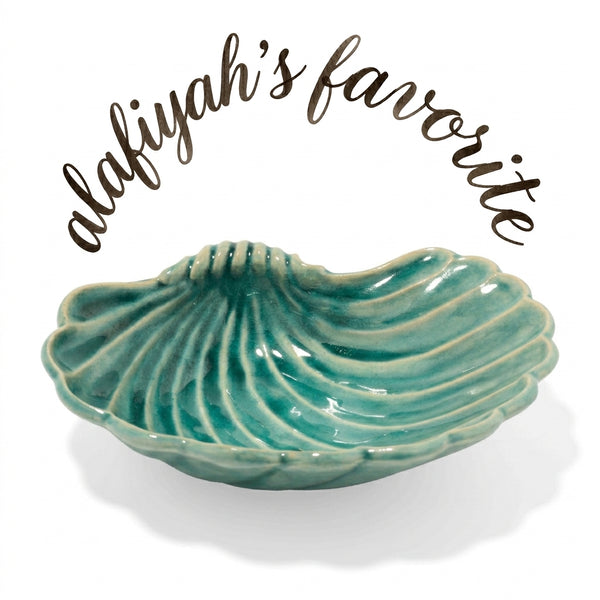 A small shell-shaped ceramic dish in a glossy teal green glaze, shown on a white background with the text 'alafiyah's favorite' arched above it.