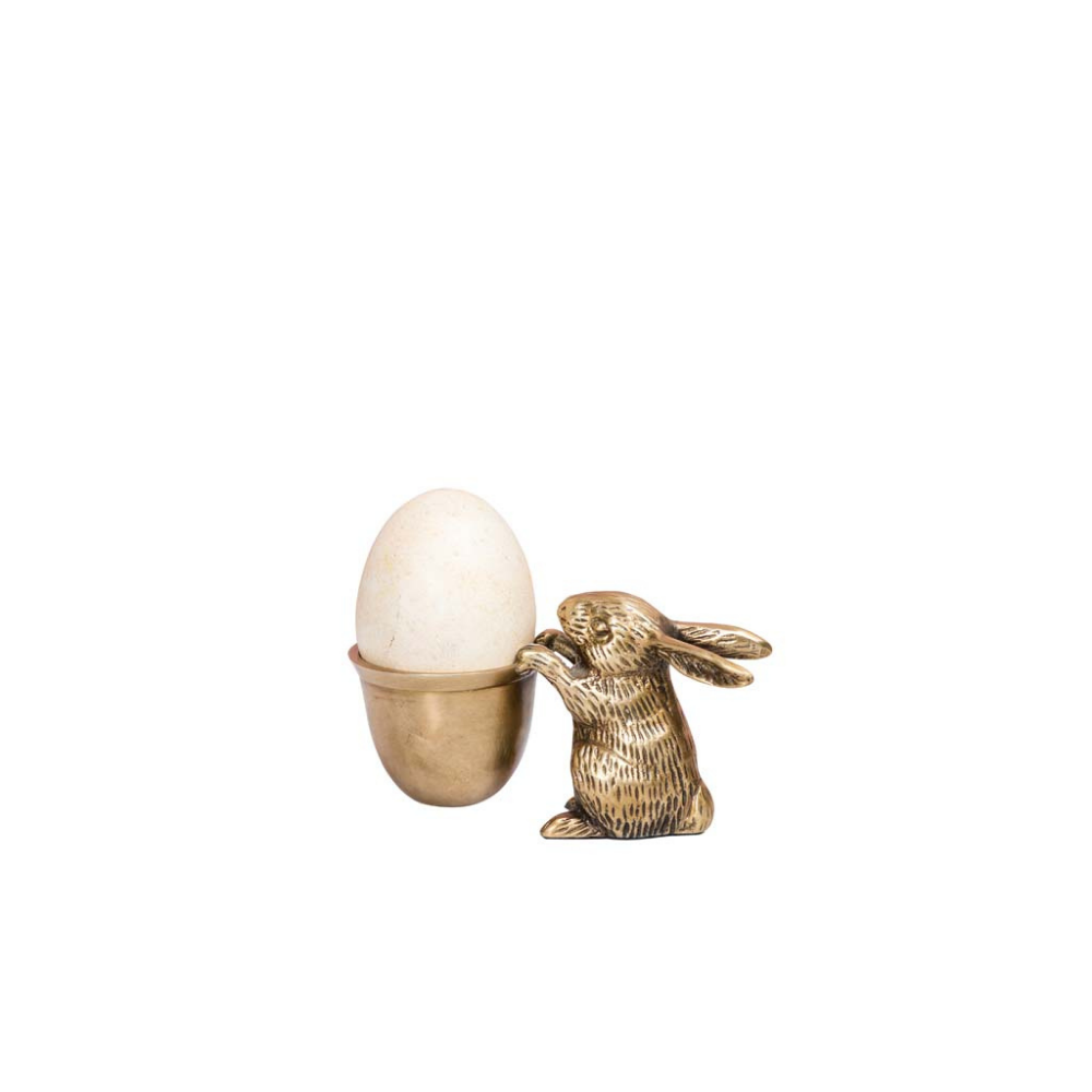 A decorative brass egg holder featuring a small rabbit figurine next to a cup-shaped holder containing a white egg, set against a plain white background.