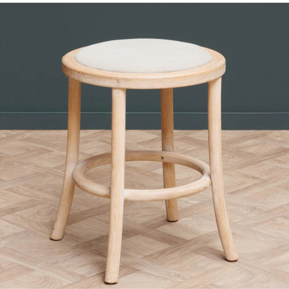 A light natural wood stool with a round off-white upholstered seat, set against a dark gray wall and a light wood herringbone floor.