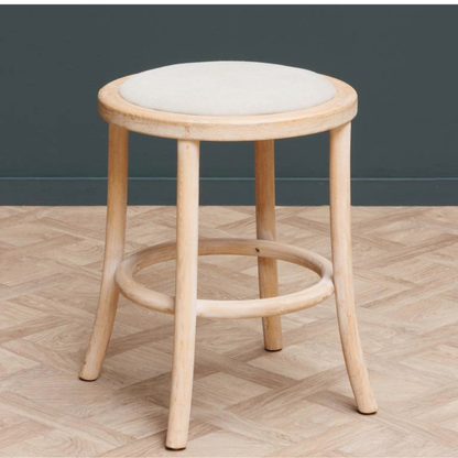 A light natural wood stool with a round off-white upholstered seat, set against a dark gray wall and a light wood herringbone floor.