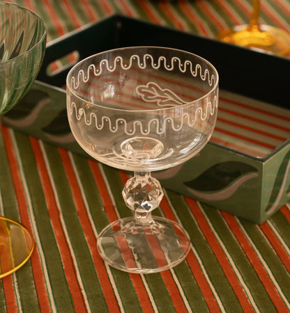 Vintage-style etched glass coupe with faceted stem, on striped tablecloth with green tray in the background.