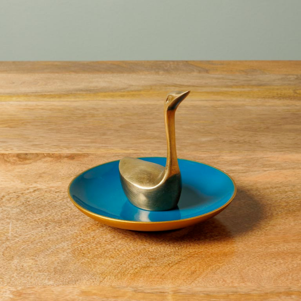 A small decorative object shaped like a goose standing on a round blue saucer with a gold rim, placed on a wooden tabletop against a soft gray background.