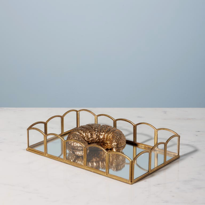 A rectangular gold-framed tray with mirrored glass panels and arched top details, holding a textured metallic gold decorative object, set on a white marble surface against a light blue background.