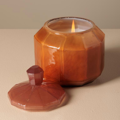 A faceted amber glass jar candle with a matching lid placed beside it on a neutral beige surface. The candle is lit, with the flame visible inside the glass container.