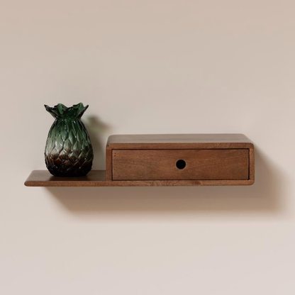 A floating wooden wall shelf with a single drawer, shown mounted on a beige wall. A green textured pineapple-shaped vase sits on the shelf to the left.