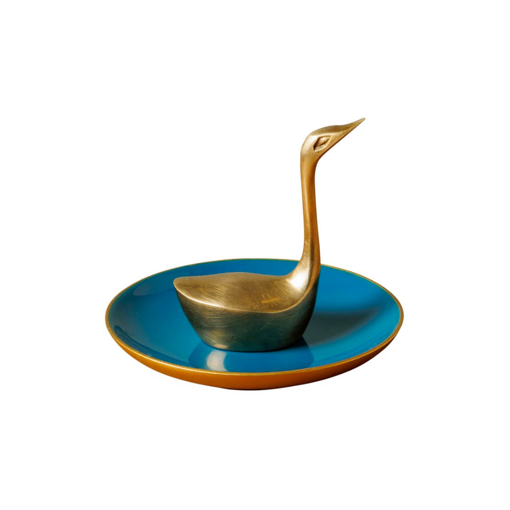 A decorative brass swan-shaped spoon resting on a round turquoise blue plate with a gold rim, set against a plain white background.