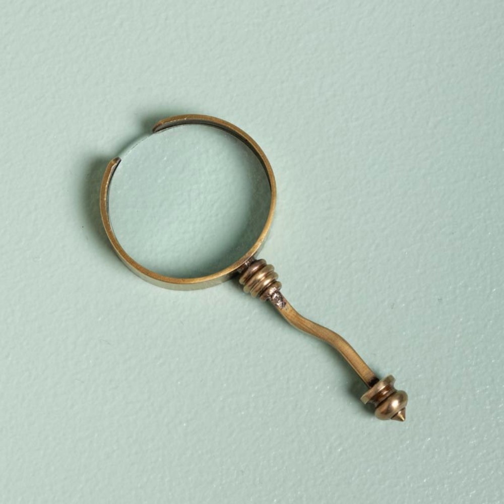 A vintage-style brass magnifying glass with a circular lens and a pointed handle, resting on a textured light blue-green background.
