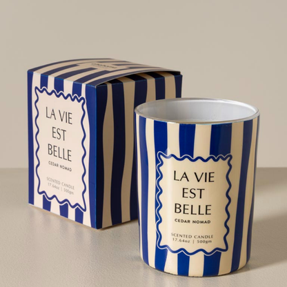 A scented candle in a cylindrical tin with blue and cream vertical stripes, shown next to its matching striped box. The label reads 'LA VIE EST BELLE' and 'CEDAR NOMAD'.
