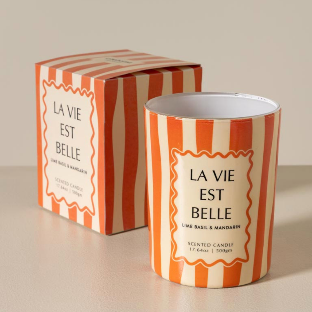A scented candle in a white tin with orange and cream vertical stripes stands in front of its matching striped box. The front label reads 'LA VIE EST BELLE' and 'LIME BASIL & MANDARIN'.