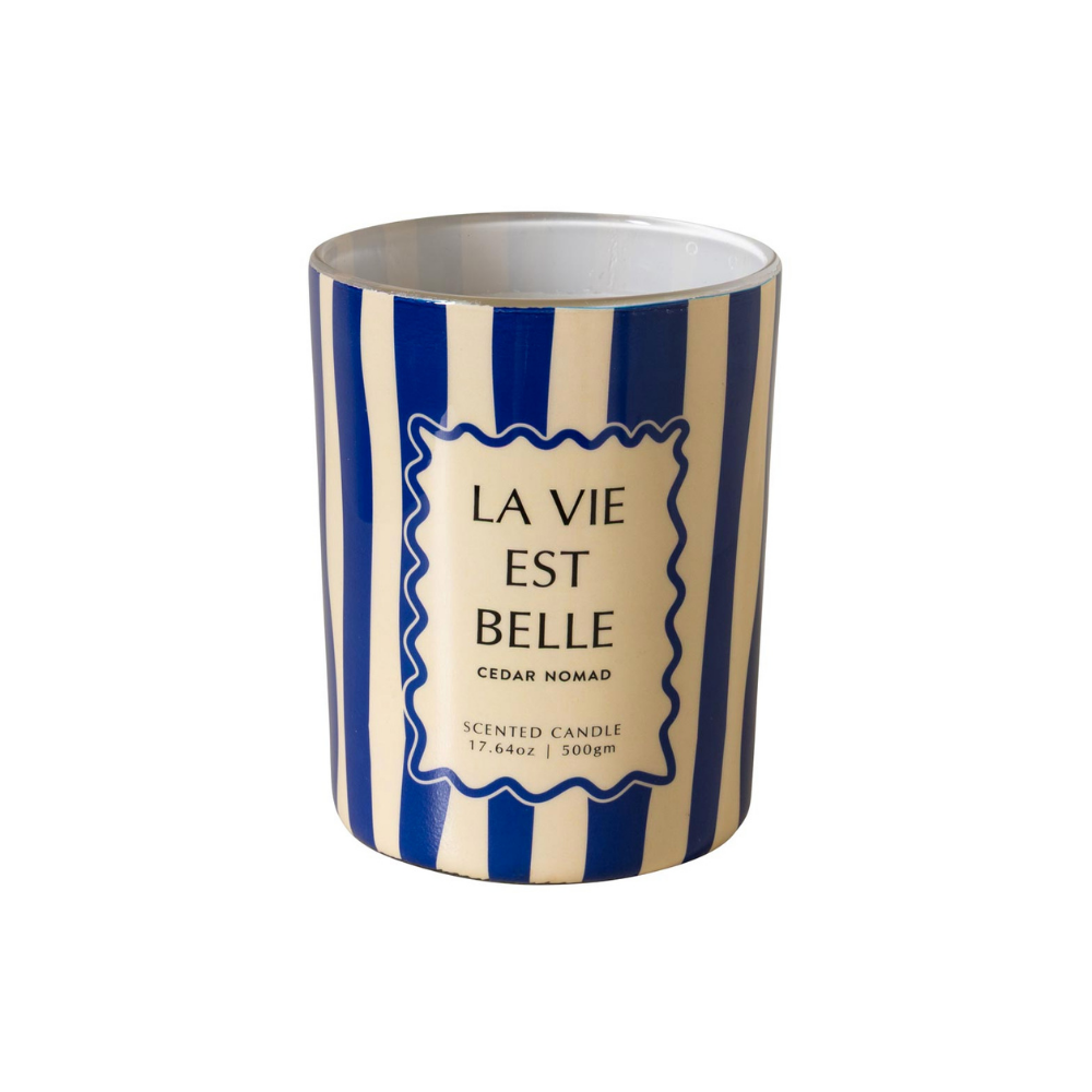 A scented candle in a cylindrical tin with a blue and cream vertical stripe pattern. The front features a cream-colored label with a wavy border and the French text 'LA VIE EST BELLE' and 'CEDAR NOMAD'.