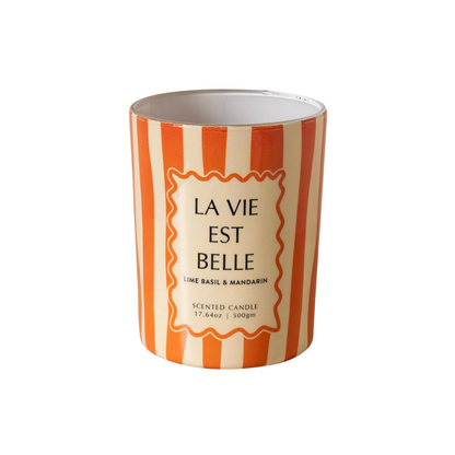 A cylindrical scented candle with a white and orange striped design, featuring a central cream-colored label with the French text 'LA VIE EST BELLE' and smaller scent details 'LIME BASIL & MANDARIN'.