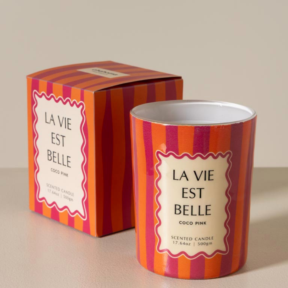 A scented candle in a striped metal tin next to its matching retail box. The packaging features bold vertical stripes in orange, pink, and red with a cream-colored label that reads 'LA VIE EST BELLE' and 'COCO PINK'.