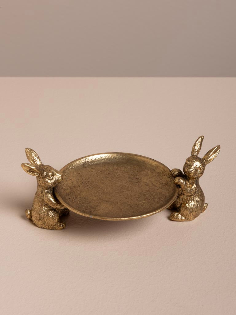 A decorative gold-toned metal tray with two rabbit figurines supporting its ends, photographed on a plain beige surface against a neutral beige background.