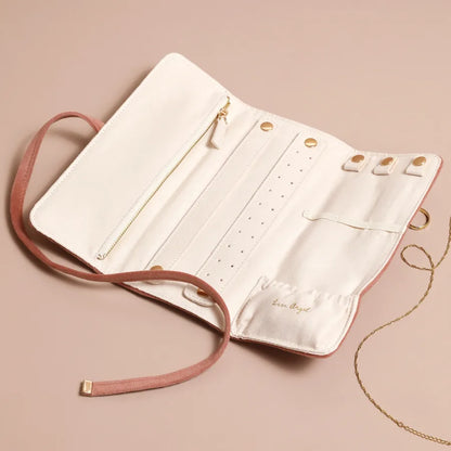An off-white faux leather jewelry organizer roll with a dusty pink faux suede strap and gold-tone hardware, shown open on a solid beige background with a gold chain draped over it.