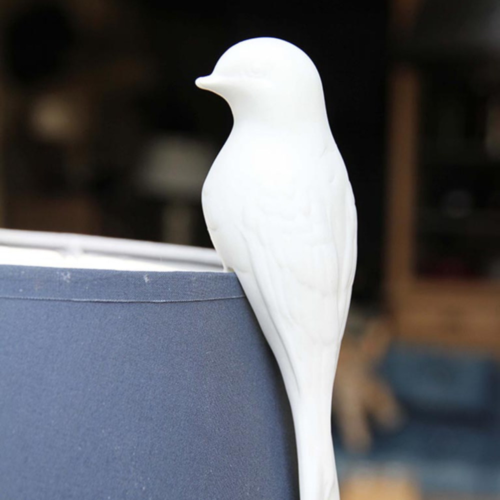 A white ceramic bird figurine perched on the edge of a dark blue lampshade, set against a softly blurred indoor background.