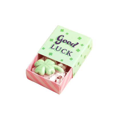 A small St. Patrick's Day themed matchbox gift box with a light green lid that reads 'Good Luck' in purple lettering, decorated with green shamrock motifs.