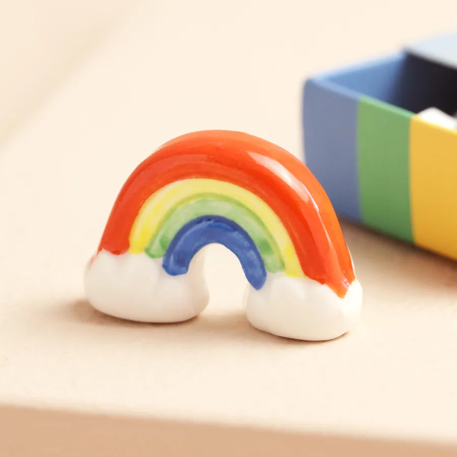 Colorful rainbow-shaped ceramic figurine on a beige surface with a blurred colorful box in the background.