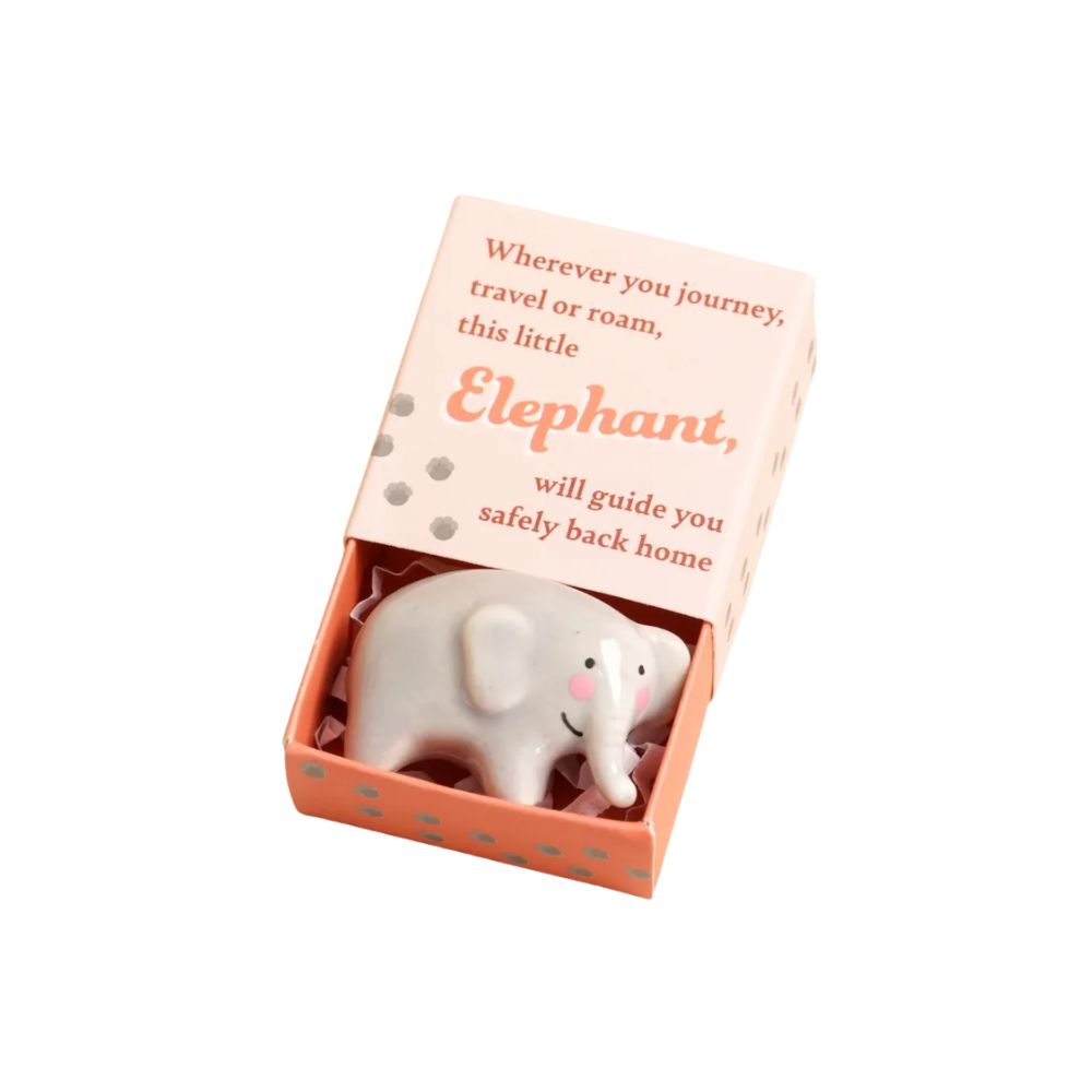 A small white elephant figurine inside an open peach-colored gift matchbox with printed text on the lid, photographed on a plain white background.