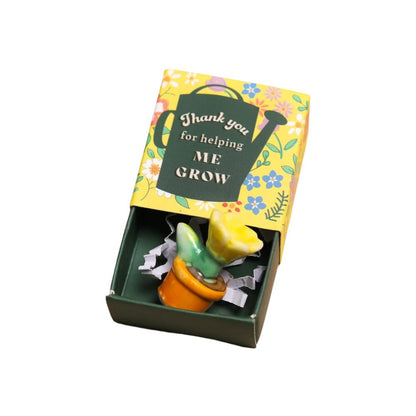 A small ceramic plant pot figurine with a yellow flower and green leaves, presented inside a small dark green gift matchbox with a floral yellow lid that reads 'Thank you for helping ME GROW'.
