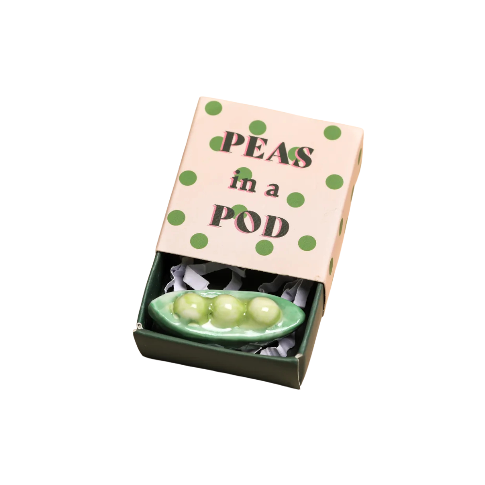 A ceramic figurine of green peas in a pod, displayed inside its open decorative gift matchbox with a pink lid featuring green polka dots and the text 'PEAS in a POD'.