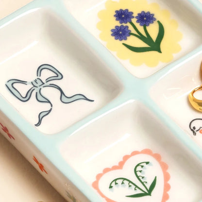 Sectioned ceramic trinket tray with bow, cornflower and heart illustrations, holding gold rings.
