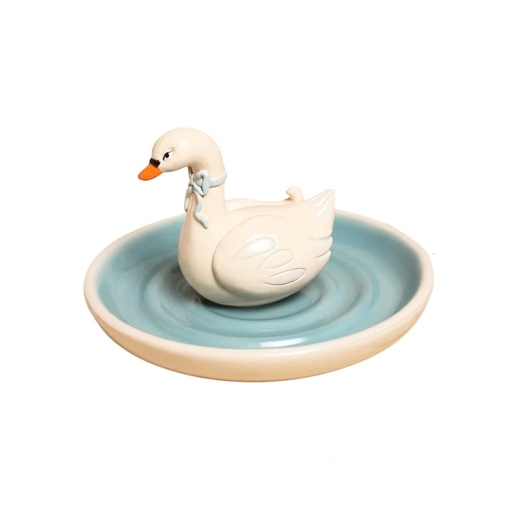 A ceramic swan figurine sitting on a shallow round dish. The swan is cream-colored with a small orange beak and a light blue ribbon around its neck, while the dish is light blue with a cream-colored rim.
