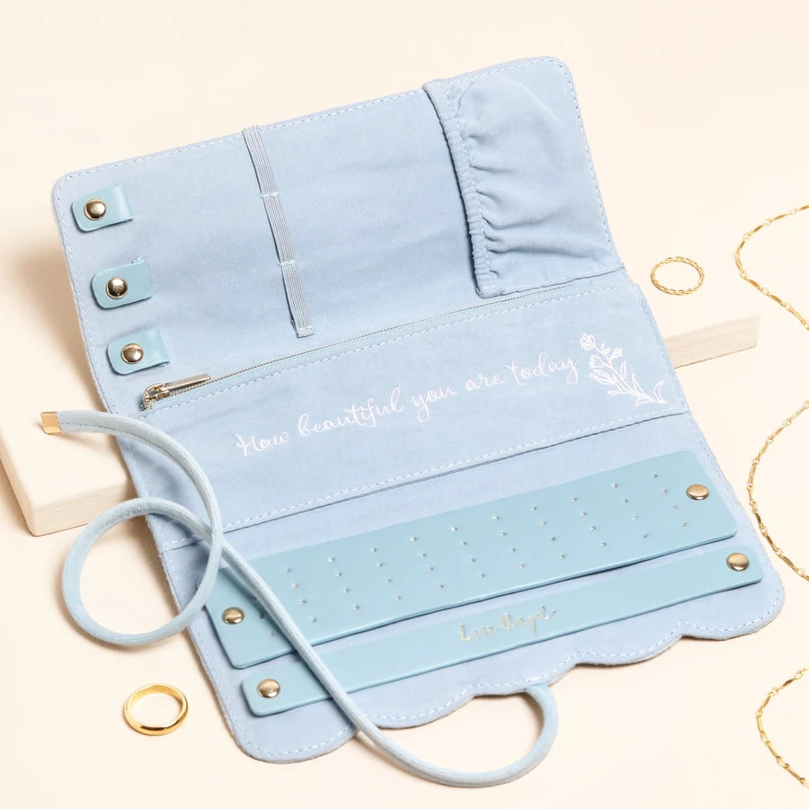 A light blue velvet jewelry organizer roll-up pouch with gold-tone hardware, shown open on a cream background with decorative gold rings and white blocks.