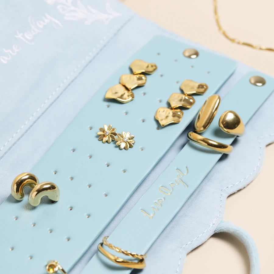 A close-up of gold-toned jewelry displayed on a light blue velvet jewelry organizer. The organizer features multiple slots holding earrings and rings, including heart-shaped studs, flower-shaped studs, crescent earrings, and ring-shaped earrings.