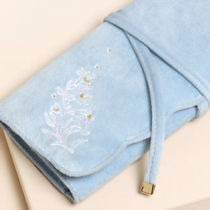 A light blue plush velvet wrap blanket with white floral embroidery and small gold bead accents, folded neatly on a beige background.