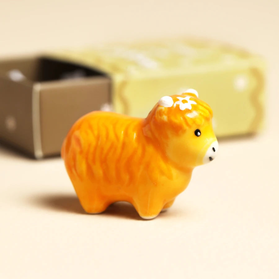 A small glossy ceramic animal figurine shaped like a fluffy yellow-orange sheep or cow, standing in front of a softly blurred pale green box on a warm beige background.