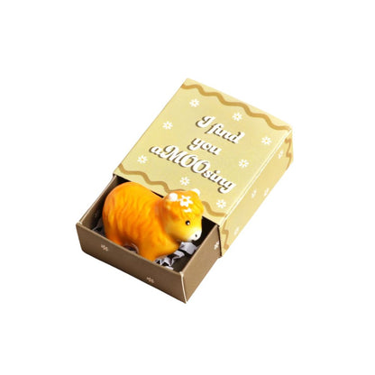 An orange ceramic cat figurine nestled inside an open decorative matchbox with a gold lid that reads 'I found you a MOOting'.