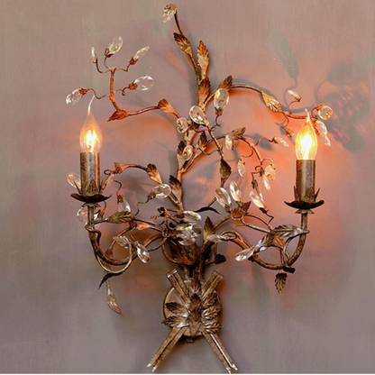 A decorative two-light wall sconce with an ornate metal branch and leaf design, mounted on a wall. The fixture features two candle-style bulbs emitting a warm glow, with clear faceted crystal-like leaf accents attached to the branch structure.