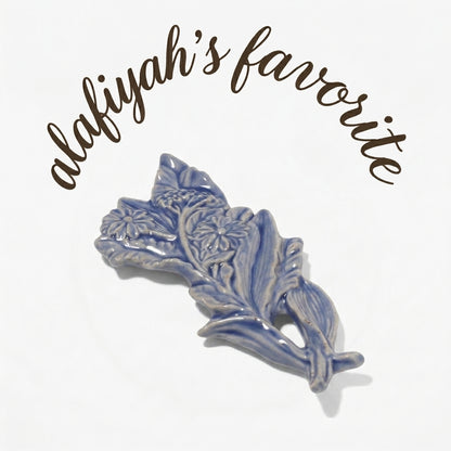 A blue and white glazed ceramic floral ornament shaped like leaves and flowers, displayed on a clean white background with the text 'alafiyah's favorite' written in brown script above it.