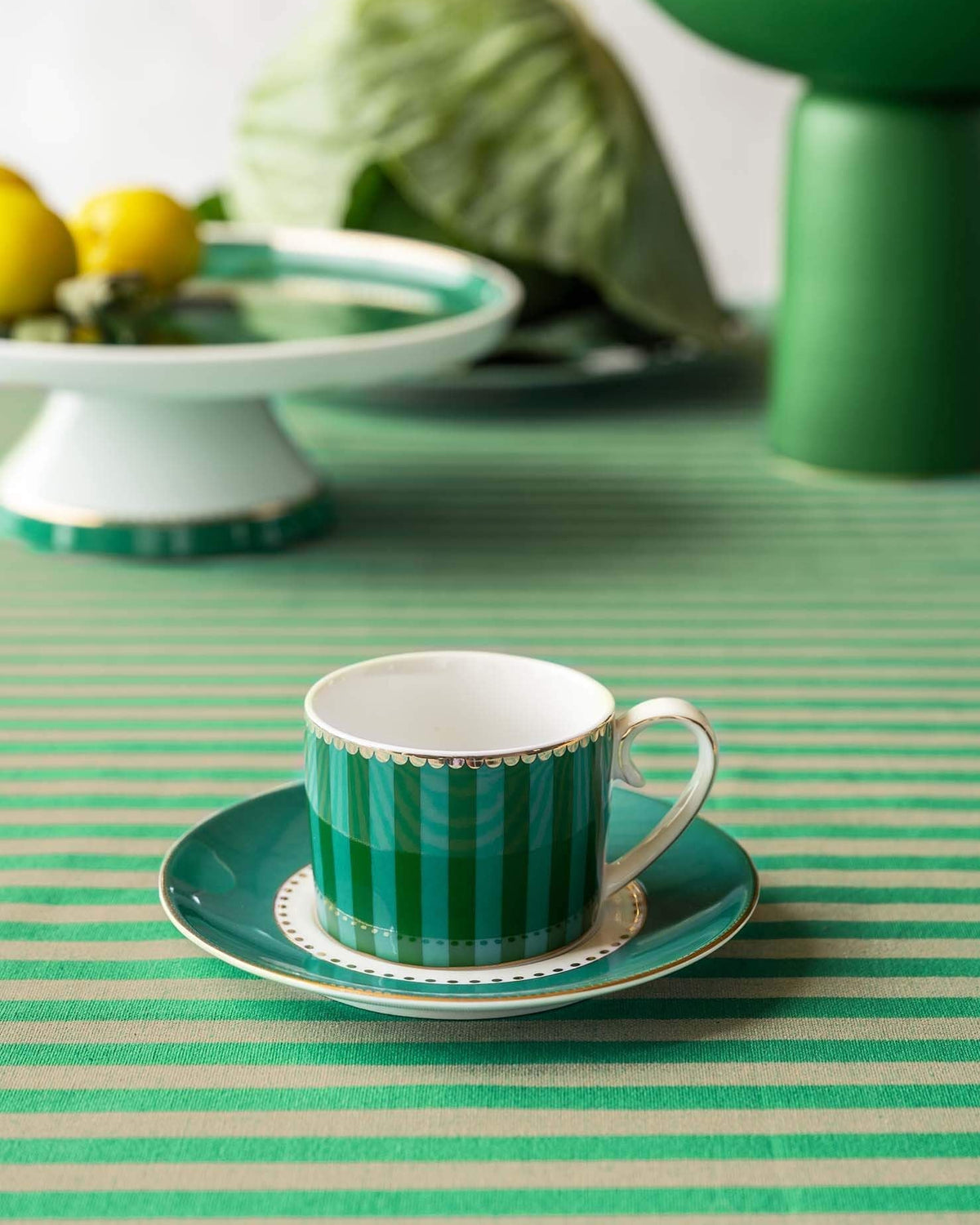 Green striped teacup and saucer on a green and white striped tablecloth with a cake stand in the background.