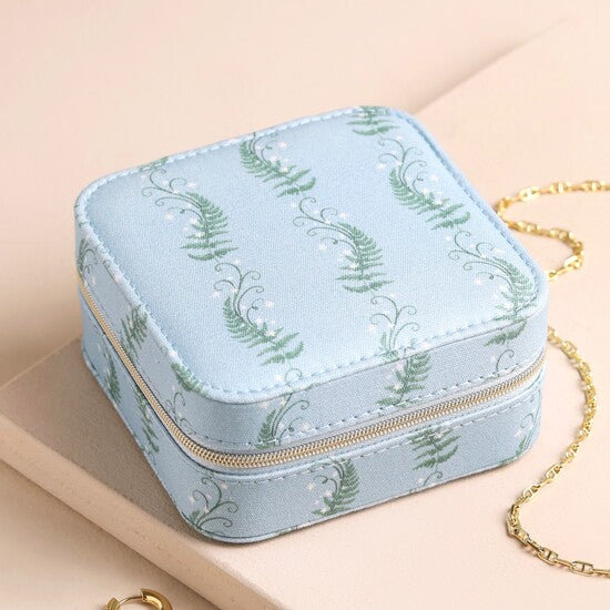 A square light blue jewelry box with a green fern pattern and a gold zipper, styled on a beige surface with a gold chain nearby.