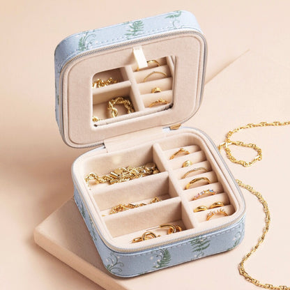 An open light blue jewelry organizer case with a green botanical pattern, filled with gold rings and necklaces, set against a warm beige background.