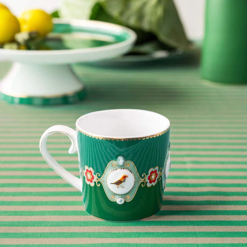 A green ceramic coffee mug with a white handle and interior, decorated with a central white oval medallion featuring a small bird and floral details, set on a green and white striped tablecloth.